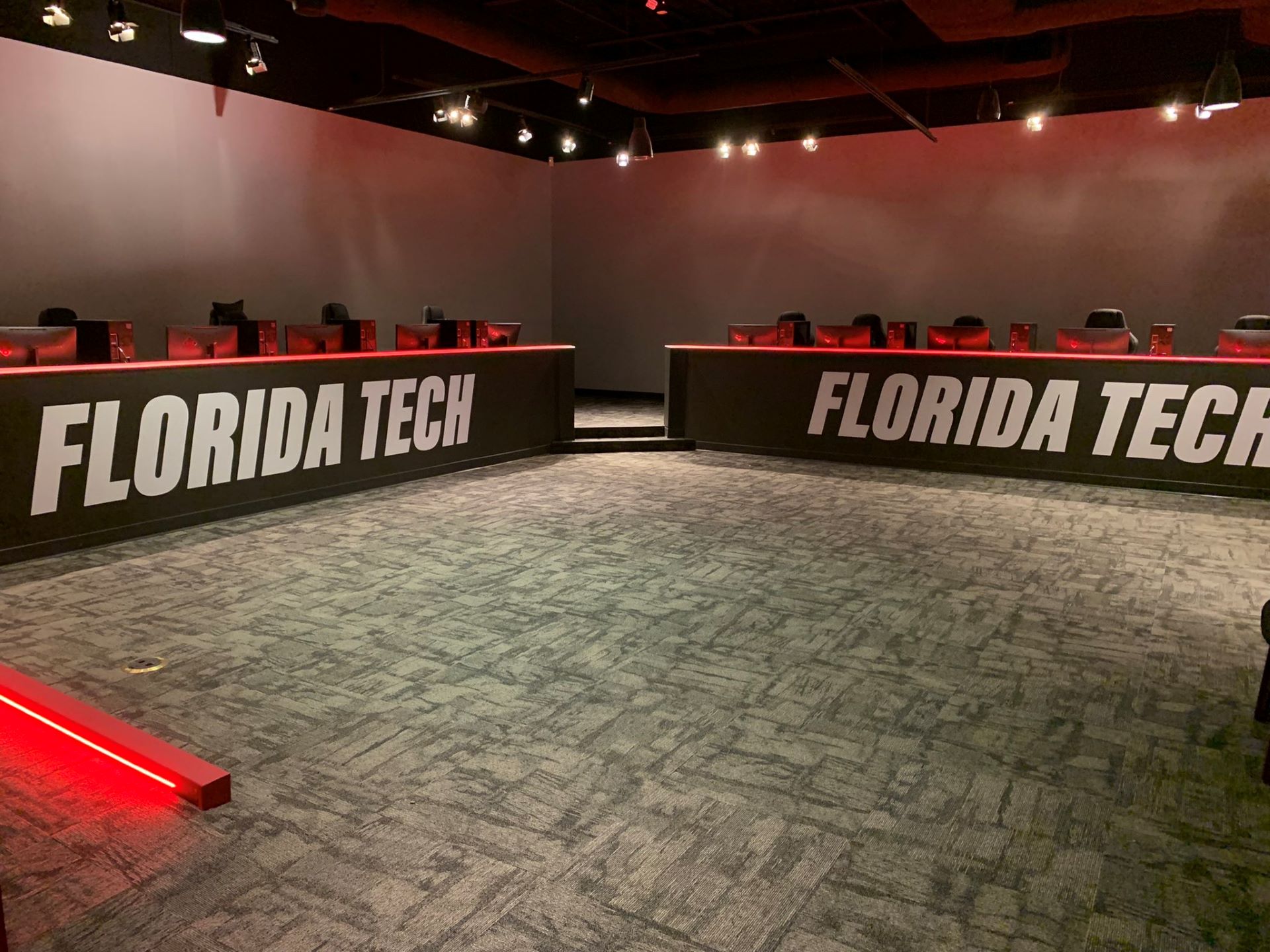 Interior of an Esports center featuring two long rows of computer stations labeled 'Florida Tech,' with multiple gaming chairs and monitors set up, illuminated by red and white lighting